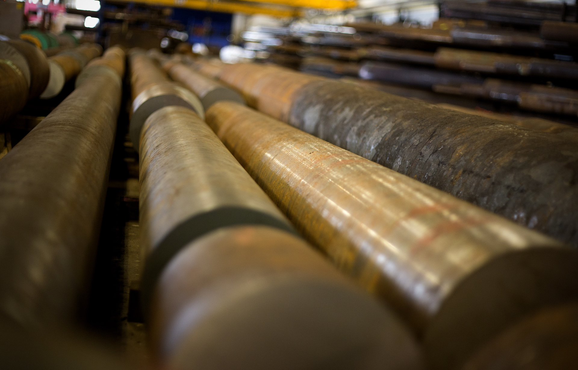 Close-up of several large, cylindrical steel pipes arranged in parallel rows inside an industrial warehouse, highlighting engineering precision, with a blurred background showing more pipes and equipment.